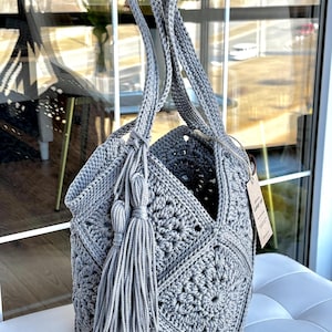 May include: A grey crochet tote bag constructed from multiple square motifs featuring a central floral pattern. The bag includes two long shoulder straps and a decorative tassel accent attached to the side. A small rectangular paper tag is attached.