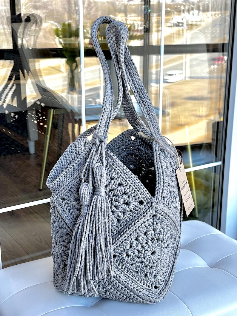 May include: A grey crochet tote bag constructed from multiple square motifs featuring a central floral pattern. The bag includes two long shoulder straps and a decorative tassel accent attached to the side. A small rectangular paper tag is attached.
