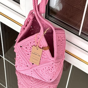 May include: A bright pink crochet bag constructed from square motifs with a single top handle. A brown paper tag is attached to the bag with string, featuring the text "CROCHET BAG BY Lunar cat" and "Thank you for purchasing".