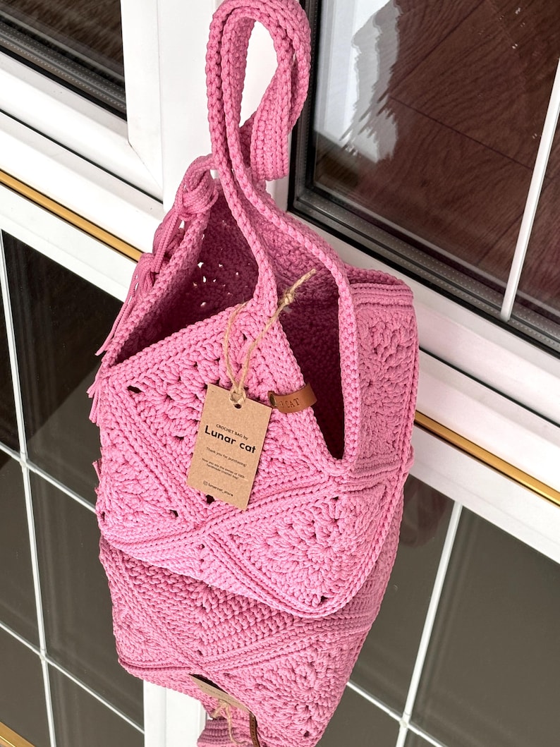 May include: A bright pink crochet bag constructed from square motifs with a single top handle. A brown paper tag is attached to the bag with string, featuring the text "CROCHET BAG BY Lunar cat" and "Thank you for purchasing".