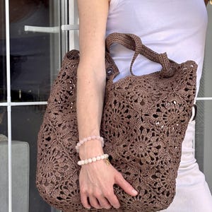 Puede incluir: Un bolso tote de ganchillo marrón que presenta un patrón de motivo floral repetitivo en todo el cuerpo. El bolso está construido con una serie de elementos de hilo circulares interconectados, creando un diseño calado con dos asas de ganchillo marrones a juego.