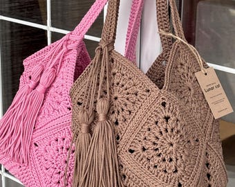 May include: Two handmade crochet tote bags, one in pink and one in brown, featuring a square motif pattern, long shoulder straps, and large matching tassels. A brown paper tag attached to the brown bag displays the text Lunar cat.