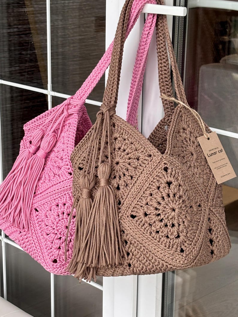 May include: Two handmade crochet tote bags, one in pink and one in brown, featuring a square motif pattern, long shoulder straps, and large matching tassels. A brown paper tag attached to the brown bag displays the text Lunar cat.