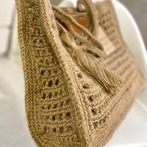 May include: A close-up of a beige crocheted tote bag with a tassel. The bag has a woven design and is made of natural fibres.