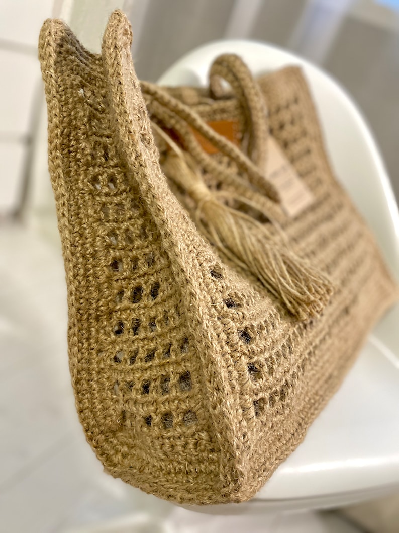 May include: A close-up of a beige crocheted tote bag with a tassel. The bag has a woven design and is made of natural fibres.