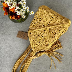 May include: Mustard yellow crochet bag with a square pattern and long straps. The bag has a tassel and a tag that reads "Lunar Cat". A vase of flowers is in the background.