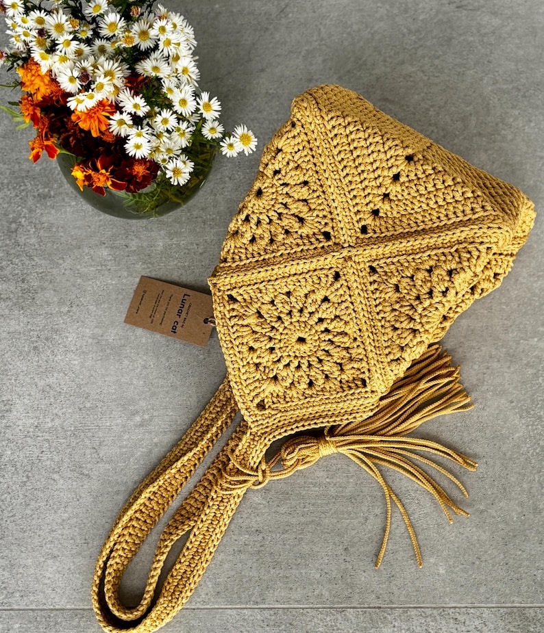 May include: Mustard yellow crochet bag with a square pattern and long straps. The bag has a tassel and a tag that reads "Lunar Cat". A vase of flowers is in the background.