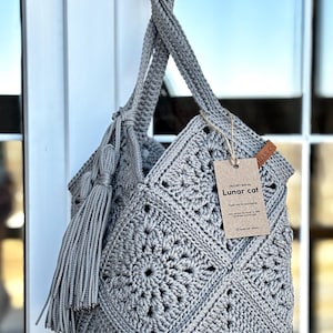 May include: A grey crochet bag constructed from square motifs with a matching tassel attached to the handle. A brown paper tag is tied to the bag with string, featuring the text "CROCHET BAG by Lunar cat" and "Thank you for purchasing!"