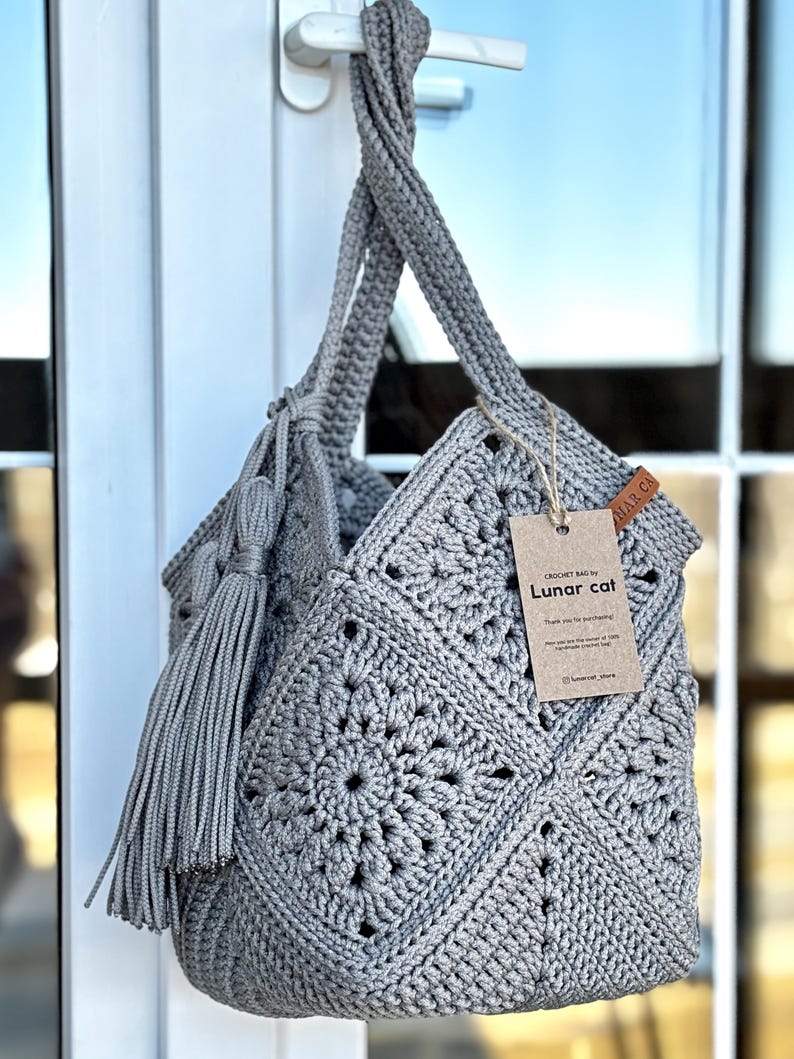 May include: A grey crochet bag constructed from square motifs with a matching tassel attached to the handle. A brown paper tag is tied to the bag with string, featuring the text "CROCHET BAG by Lunar cat" and "Thank you for purchasing!"