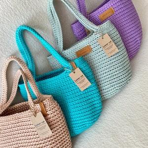 May include: Four handmade crochet tote bags in various colors: beige, turquoise, light blue, and purple. Each bag has a sturdy handle and a tag that reads "Lunar Cat". The bags are made of thick yarn and have a textured appearance.