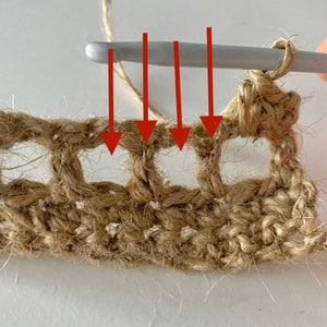 May include: Close-up of a crochet project in progress, showing a crochet hook inserted into four loops of the previous row. The hook is made of white plastic and the yarn is a natural brown colour.
