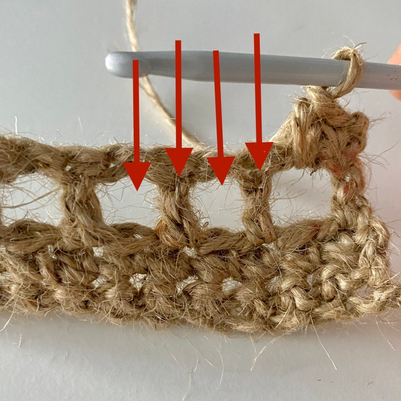 May include: Close-up of a crochet project in progress, showing a crochet hook inserted into four loops of the previous row. The hook is made of white plastic and the yarn is a natural brown colour.