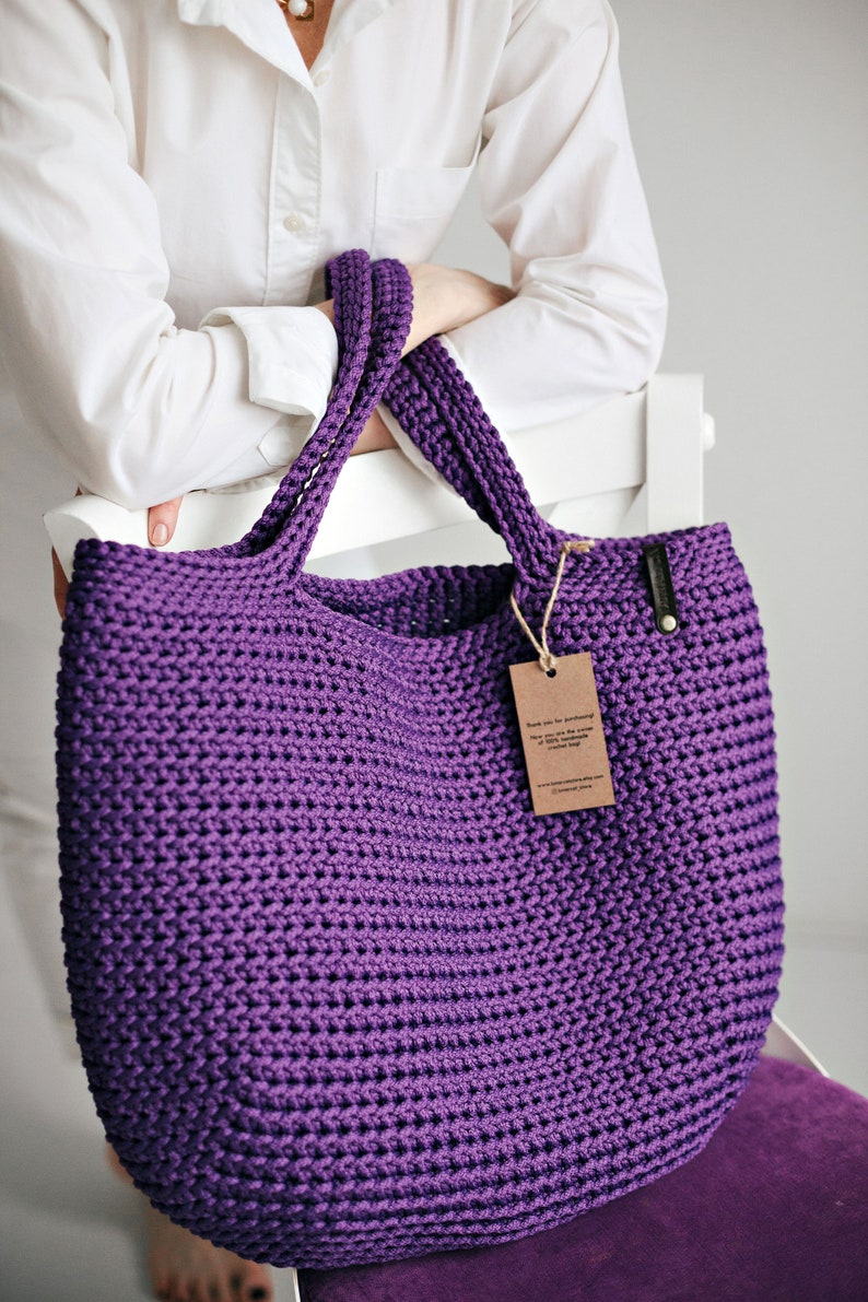 May include: A large purple crocheted tote bag with thick straps. The bag is made of a soft, textured yarn and has a simple, classic design.