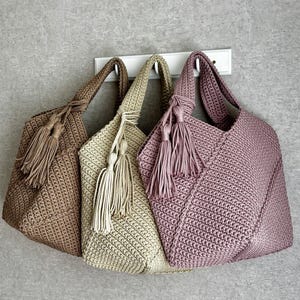 May include: Three handmade crochet tote bags in shades of brown, beige, and mauve hang on a white rack. Each bag features a matching tassel and a sturdy handle. The bags are made with a tight stitch pattern.