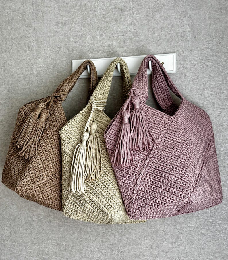 May include: Three handmade crochet tote bags in shades of brown, beige, and mauve hang on a white rack. Each bag features a matching tassel and a sturdy handle. The bags are made with a tight stitch pattern.