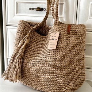 Puede incluir: Un bolso tote grande tejido en color beige natural. El bolso presenta asas trenzadas gruesas y una borla larga en un lado. Una etiqueta con el texto "XXL Size Lunar cat" est&aacute; sujeta al bolso.