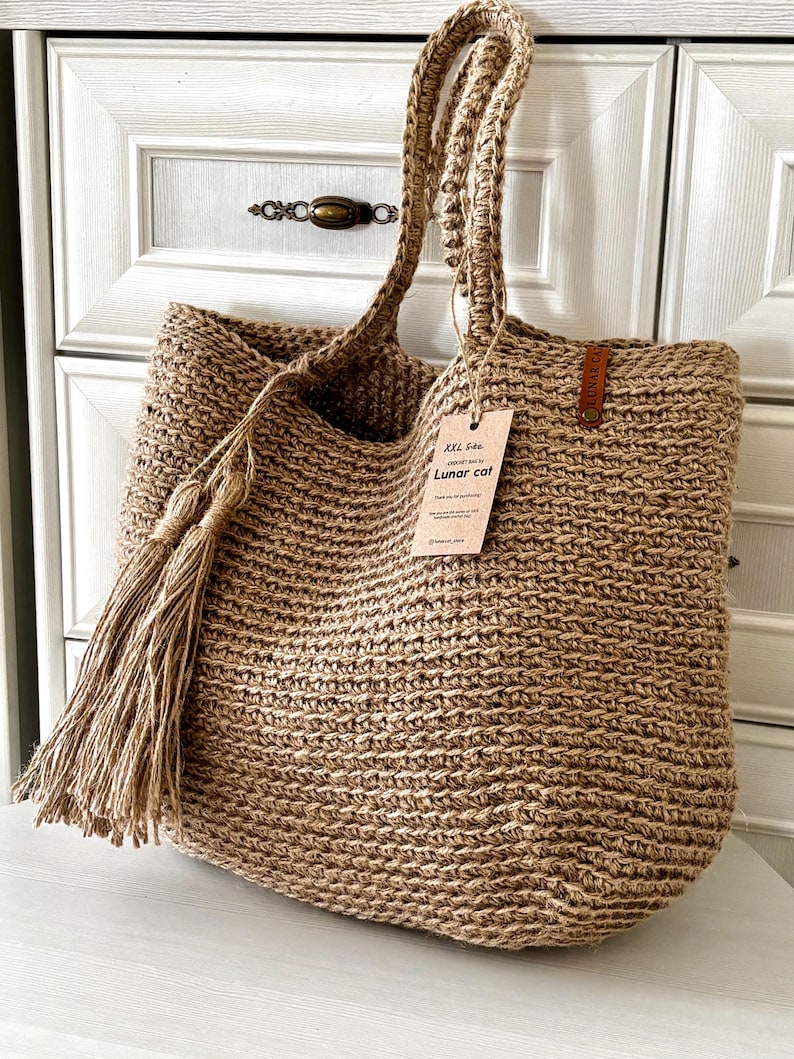 Puede incluir: Un bolso tote grande tejido en color beige natural. El bolso presenta asas trenzadas gruesas y una borla larga en un lado. Una etiqueta con el texto "XXL Size Lunar cat" est&aacute; sujeta al bolso.