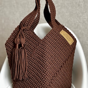 May include: A brown macrame tote bag with a knotted handle and a decorative tassel. The bag features a woven texture and a small rectangular tag with text. The bag is sitting on a white chair.