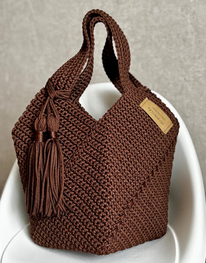 May include: A brown macrame tote bag with a knotted handle and a decorative tassel. The bag features a woven texture and a small rectangular tag with text. The bag is sitting on a white chair.