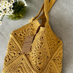 May include: Mustard yellow crochet tote bag with a geometric pattern. The bag has long straps and a tag that reads "Lunar cat". The bag is handmade and has a textured appearance. A vase of white flowers is in the background.