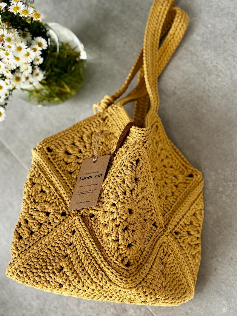 May include: Mustard yellow crochet tote bag with a geometric pattern. The bag has long straps and a tag that reads "Lunar cat". The bag is handmade and has a textured appearance. A vase of white flowers is in the background.