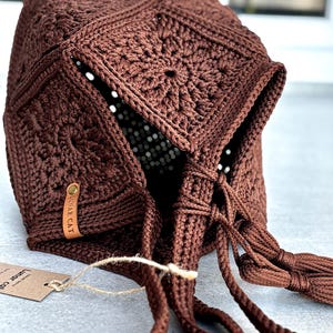 May include: A dark brown crochet bonnet constructed from multiple square motifs with a central floral pattern. The bonnet features long braided ties with tassels at the ends and a small tan leather tag embossed with the text LUNAR CAT.