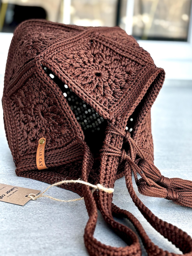 May include: A dark brown crochet bonnet constructed from multiple square motifs with a central floral pattern. The bonnet features long braided ties with tassels at the ends and a small tan leather tag embossed with the text LUNAR CAT.