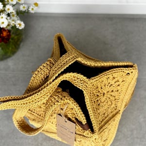 May include: Mustard yellow crochet tote bag with a square base and open top. The bag has a handle and a tag that reads "Lunar cat". The bag is made of a thick, textured yarn.