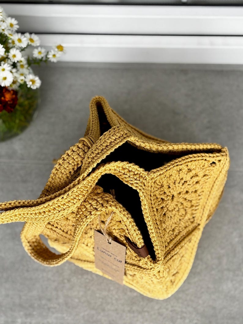 May include: Mustard yellow crochet tote bag with a square base and open top. The bag has a handle and a tag that reads "Lunar cat". The bag is made of a thick, textured yarn.