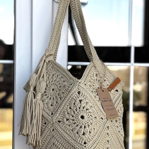 May include: A beige crochet tote bag featuring a square motif pattern, a single shoulder strap, and two long decorative tassels attached to the side. A brown leather tag labeled Lunar Cat is attached to the bag with a brown string.