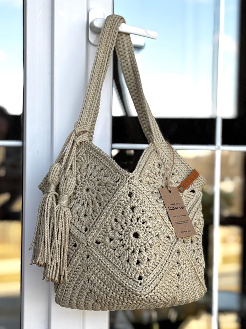 May include: A beige crochet tote bag featuring a square motif pattern, a single shoulder strap, and two long decorative tassels attached to the side. A brown leather tag labeled Lunar Cat is attached to the bag with a brown string.