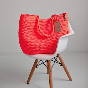 May include: A bright orange, hand-crocheted tote bag with two handles, resting on a white chair with wooden legs. The bag has a tag attached. The bag is made of a thick, textured yarn.