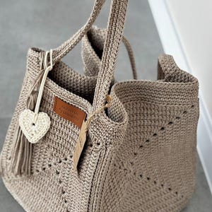 May include: A beige crochet tote bag with two handles. The bag features a heart-shaped charm and a leather tag that reads "LUNAR CAT". The bag is made of a thick yarn and has a textured appearance. The bag is open at the top.