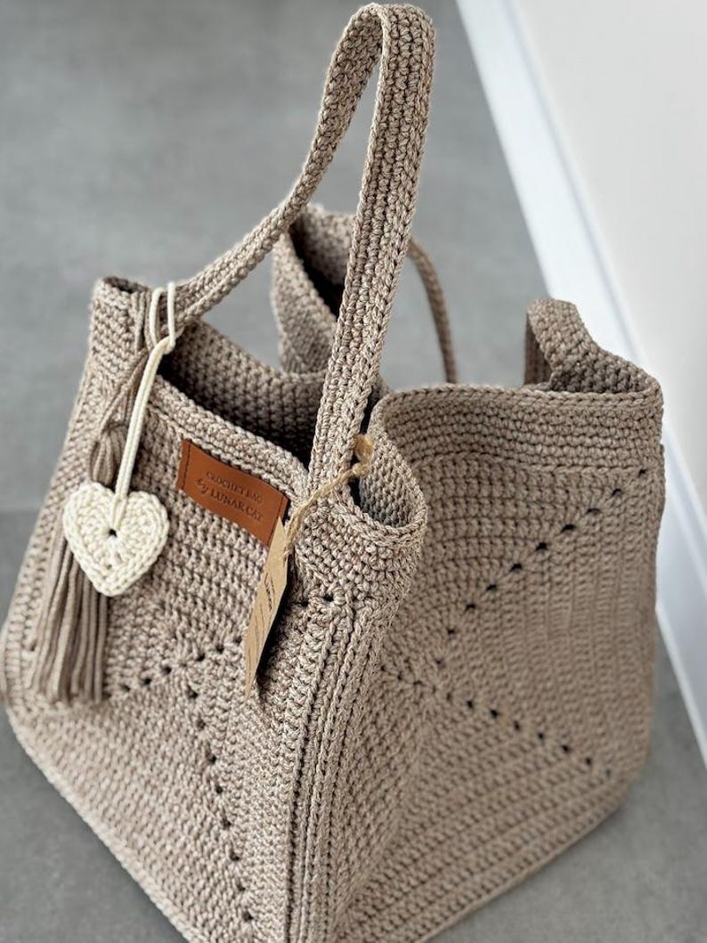 May include: A beige crochet tote bag with two handles. The bag features a heart-shaped charm and a leather tag that reads "LUNAR CAT". The bag is made of a thick yarn and has a textured appearance. The bag is open at the top.