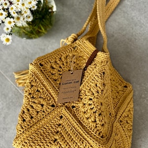 May include: Mustard yellow crochet bag with a long strap and a decorative tassel. The bag features a geometric pattern and a tag that reads "Lunar cat". The bag is made of a thick yarn and has a soft, textured appearance.