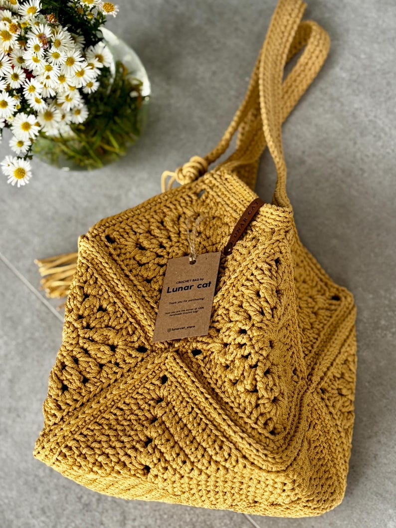 May include: Mustard yellow crochet bag with a long strap and a decorative tassel. The bag features a geometric pattern and a tag that reads "Lunar cat". The bag is made of a thick yarn and has a soft, textured appearance.