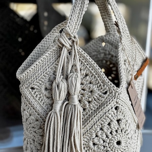 Puede incluir: Un bolso tote beige tejido a ganchillo con un patrón geométrico. El bolso tiene dos asas largas y borlas decorativas. Una pequeña etiqueta está sujeta al lateral. El bolso está hecho de un hilo grueso y texturizado.