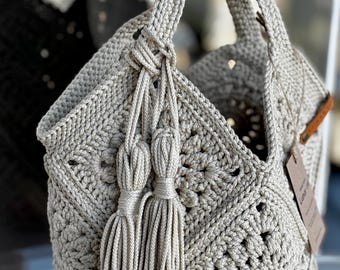 May include: A beige, crocheted tote bag with a geometric pattern. The bag features two long handles and decorative tassels. A small tag is attached to the side. The bag is made of a thick, textured yarn.