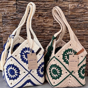 May include: Two handmade crochet bags with square motifs. One bag features cream and dark blue yarn, the other cream and dark green. Both have top handles, a side tassel, and a brown paper tag printed with "CROCHET BAG BY Lunar cat".