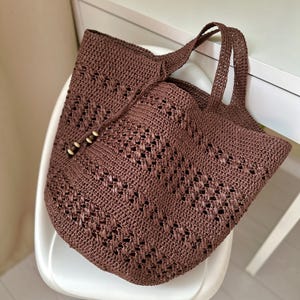 K&ouml;nnte beinhalten: Eine braune geh&auml;kelte Tragetasche mit einem gewebten Muster. Die Tasche hat zwei Henkel und ein Perlendetail.