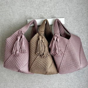 May include: Three handmade crochet tote bags in shades of mauve and tan. Each bag features a textured stitch pattern, a knotted handle, and a matching tassel. The bags are displayed against a neutral gray background, showcasing their handcrafted details.