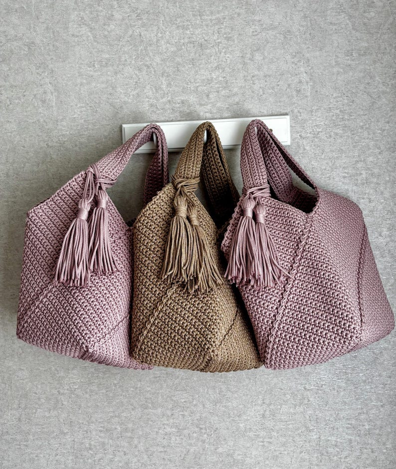 May include: Three handmade crochet tote bags in shades of mauve and tan. Each bag features a textured stitch pattern, a knotted handle, and a matching tassel. The bags are displayed against a neutral gray background, showcasing their handcrafted details.