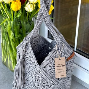 May include: A grey crochet bag constructed from square motifs with a single handle and a side tassel. A brown paper tag attached with twine displays the text "CROCHET BAG by Lunar cat" and "Thank you for purchasing". A small leather label reads "LUNAR CAT".