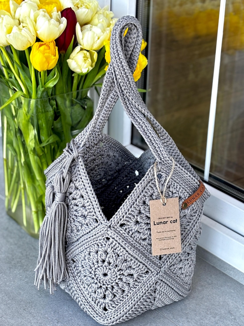May include: A grey crochet bag constructed from square motifs with a single handle and a side tassel. A brown paper tag attached with twine displays the text "CROCHET BAG by Lunar cat" and "Thank you for purchasing". A small leather label reads "LUNAR CAT".