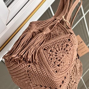 May include: A brown crochet handbag constructed from multiple square motifs with a textured pattern. The bag features two long shoulder straps and a drawstring closure adorned with two long, matching brown tassels. A small brown paper tag is attached.