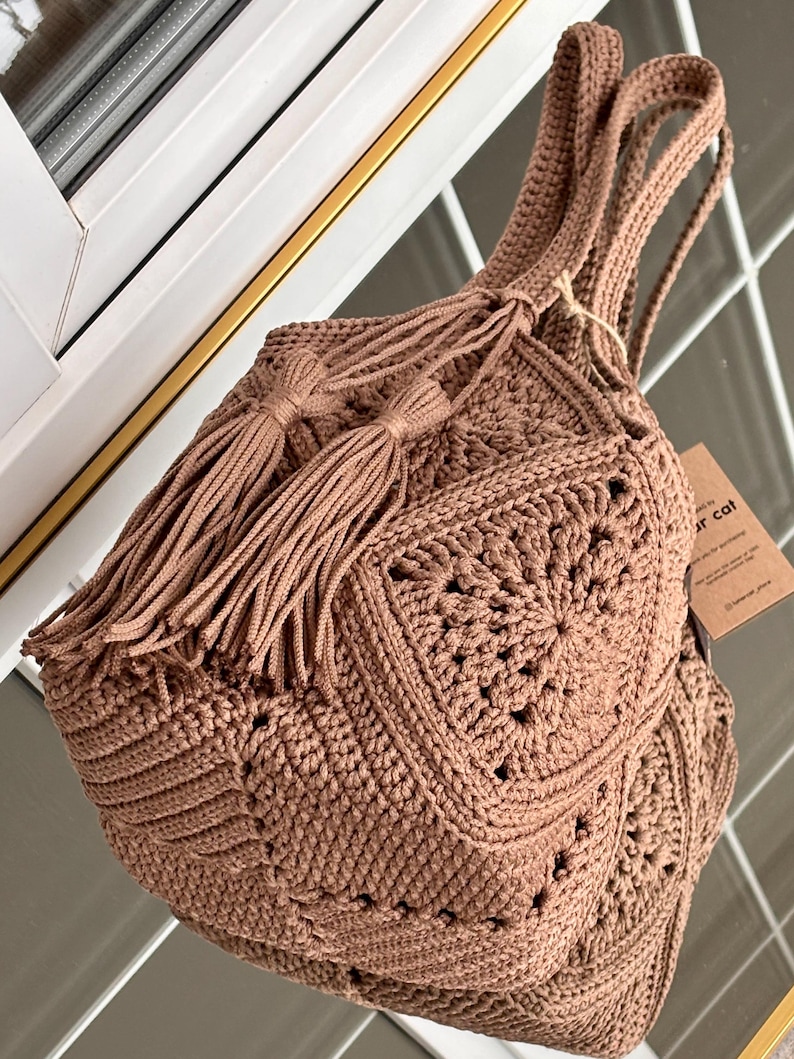 May include: A brown crochet handbag constructed from multiple square motifs with a textured pattern. The bag features two long shoulder straps and a drawstring closure adorned with two long, matching brown tassels. A small brown paper tag is attached.