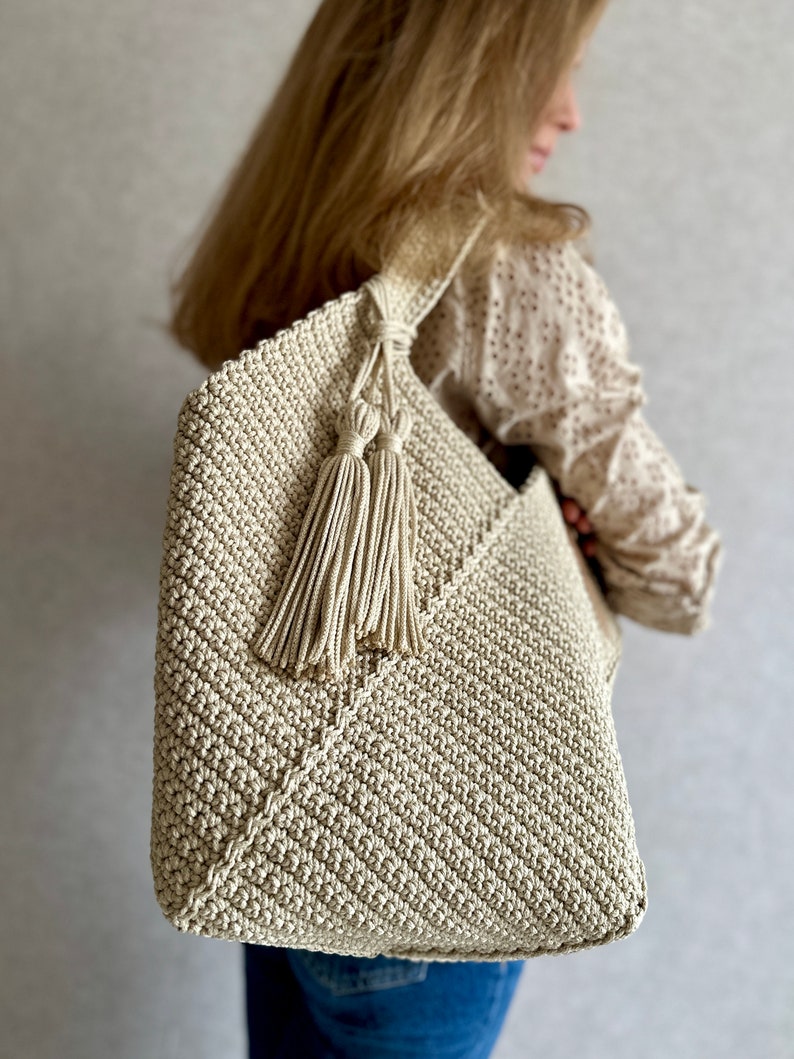 May include: A beige crocheted tote bag with a long strap and tassel accents. The bag is made of a textured, woven material.