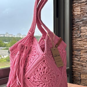 May include: A bright pink crochet tote bag featuring a square motif pattern, two long shoulder straps, and a decorative tassel hanging from the side. A brown rectangular paper tag with the text Lunar cat is attached to the bag with twine.