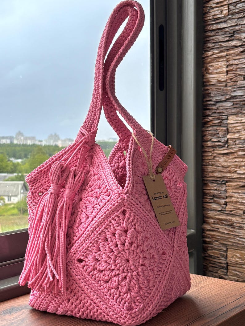 May include: A bright pink crochet tote bag featuring a square motif pattern, two long shoulder straps, and a decorative tassel hanging from the side. A brown rectangular paper tag with the text Lunar cat is attached to the bag with twine.