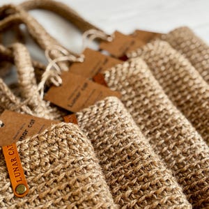 Puede incluir: Primer plano de varias bolsas de mano de yute tejido. Cada bolsa tiene un asa a juego y una etiqueta marr&oacute;n con el texto "LUNAR CAT" impreso. Las bolsas son de color marr&oacute;n natural y parecen hechas a mano.